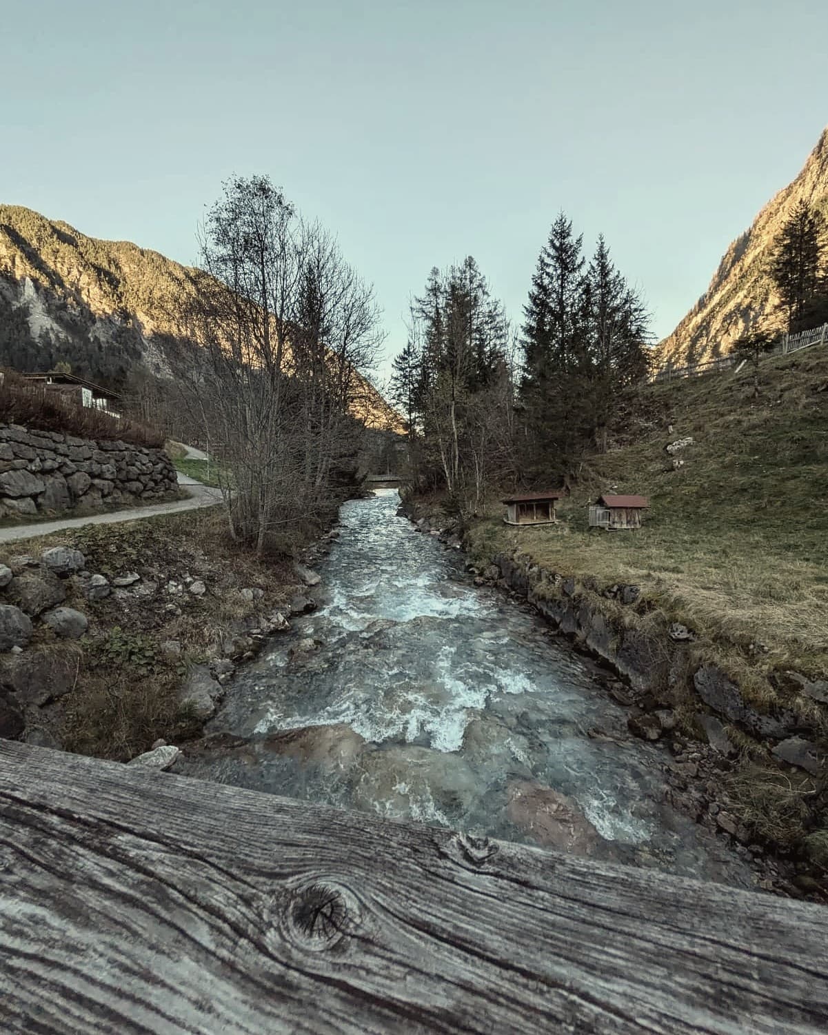 Gebirgsbach mit Steinufern, Wiese mit Heustadeln und Berge im Abendlicht